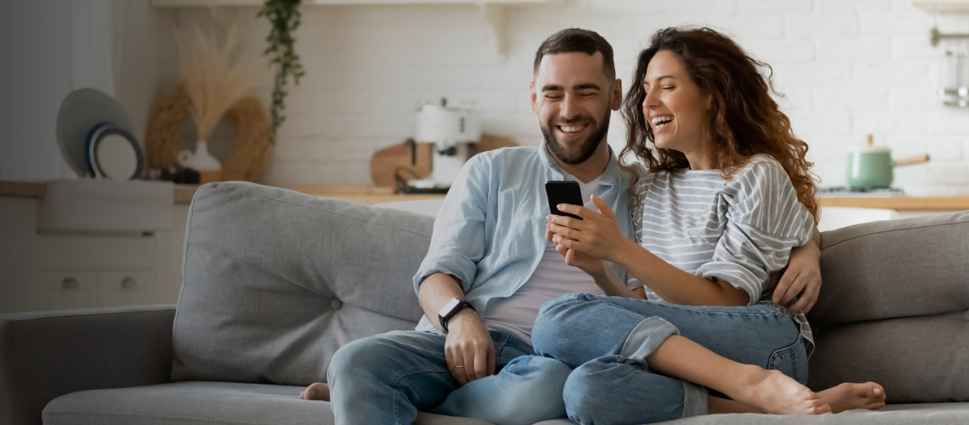 A man and a woman sit on a couch, laughing and looking at a smartphone.