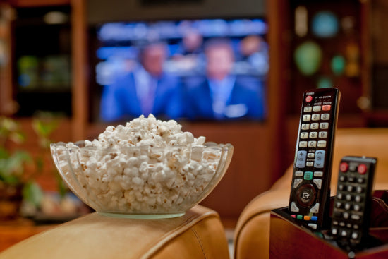 A bowl of popcorn next to tv remotes with a tv in the background